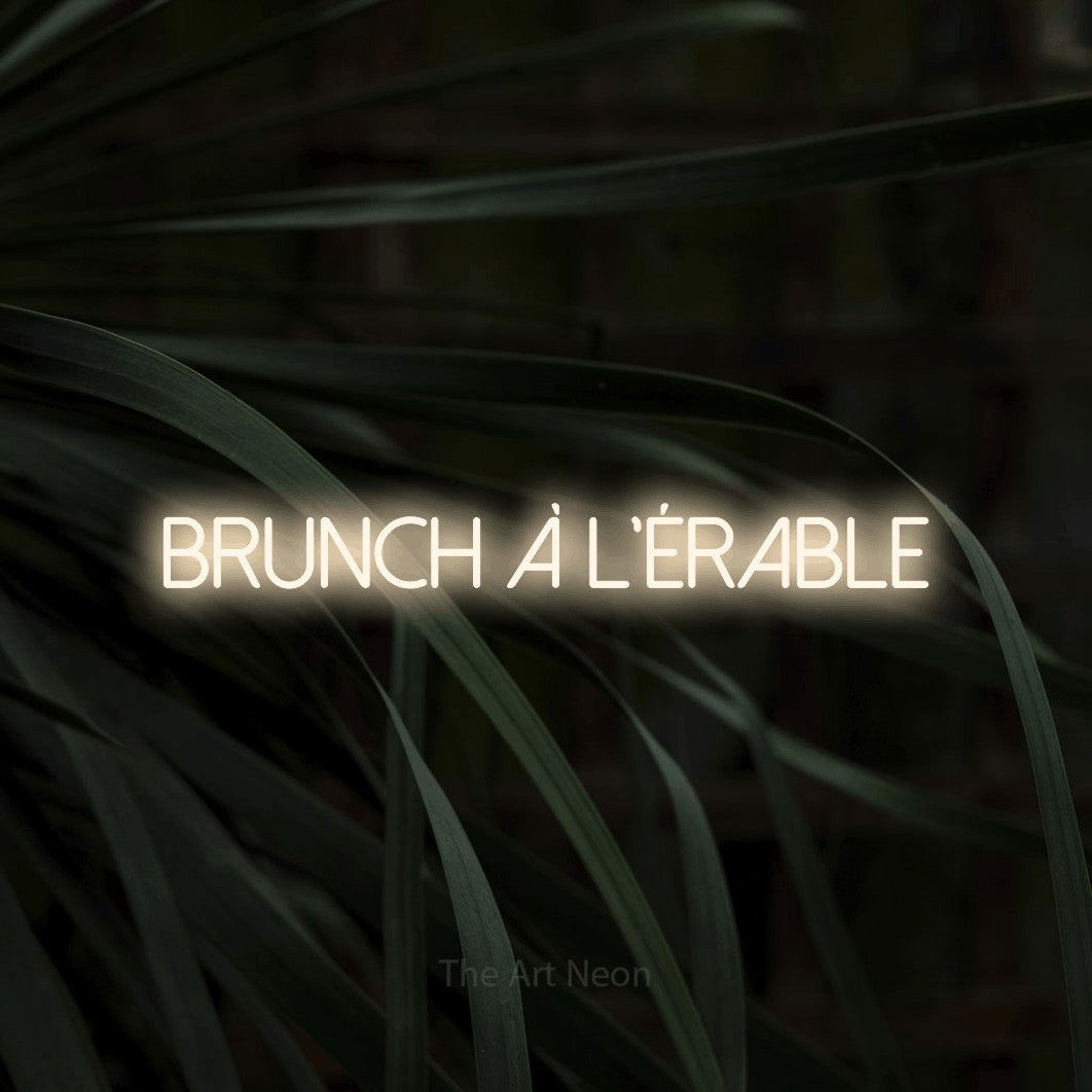 “Brunch à l'érable” LED neon sign in warm white, with thin sans-serif lettering, shown against a close-up of dark foliage.