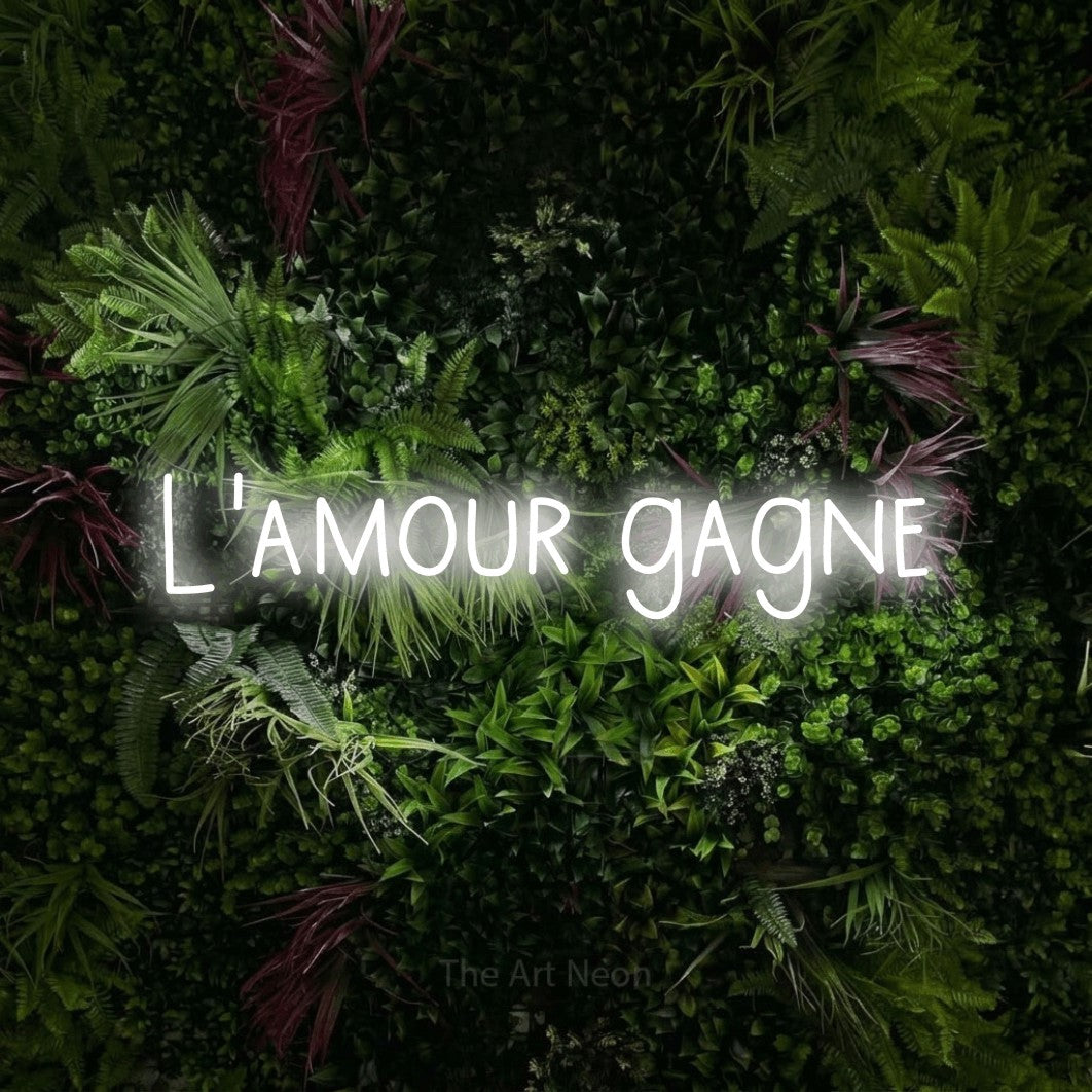 “L'amour gagne” LED neon sign in cool white, with narrow hand-drawn lettering, shown against a plant wall with pink flowers.