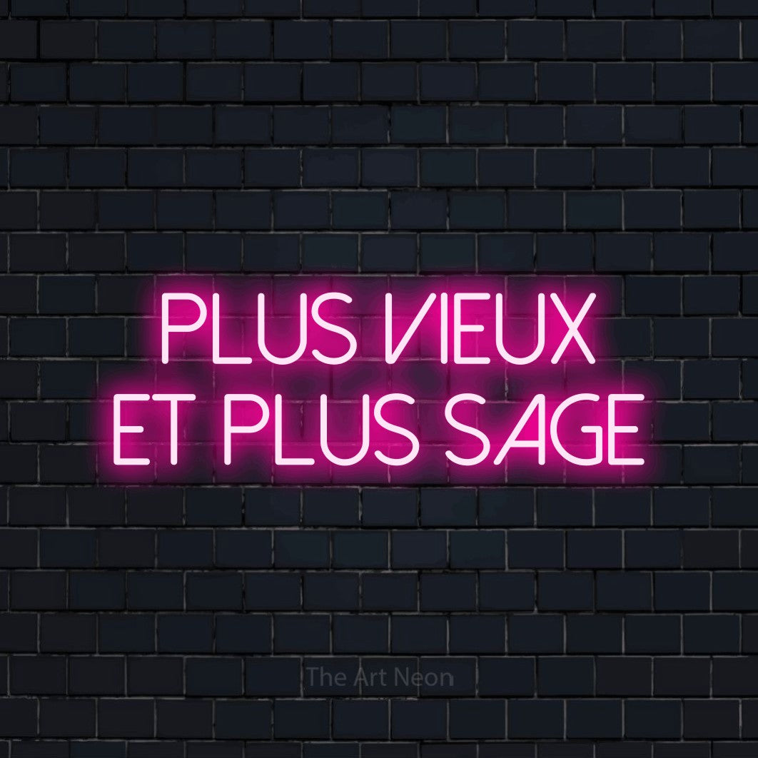 “Plus vieux et plus sage” LED neon sign in hot pink, with thin sans-serif lettering across 2 lines, shown against a dark navy brick wall.