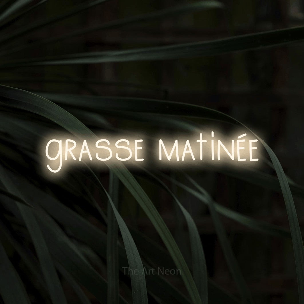 “Grasse matinée” LED neon sign in warm white, with narrow hand-drawn lettering, shown against a close-up of dark foliage.