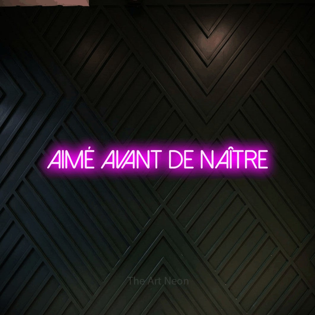 “Aimé avant de naître” LED neon sign in pink, with thin sans-serif lettering, shown against a black chevron-patterned wood wall.