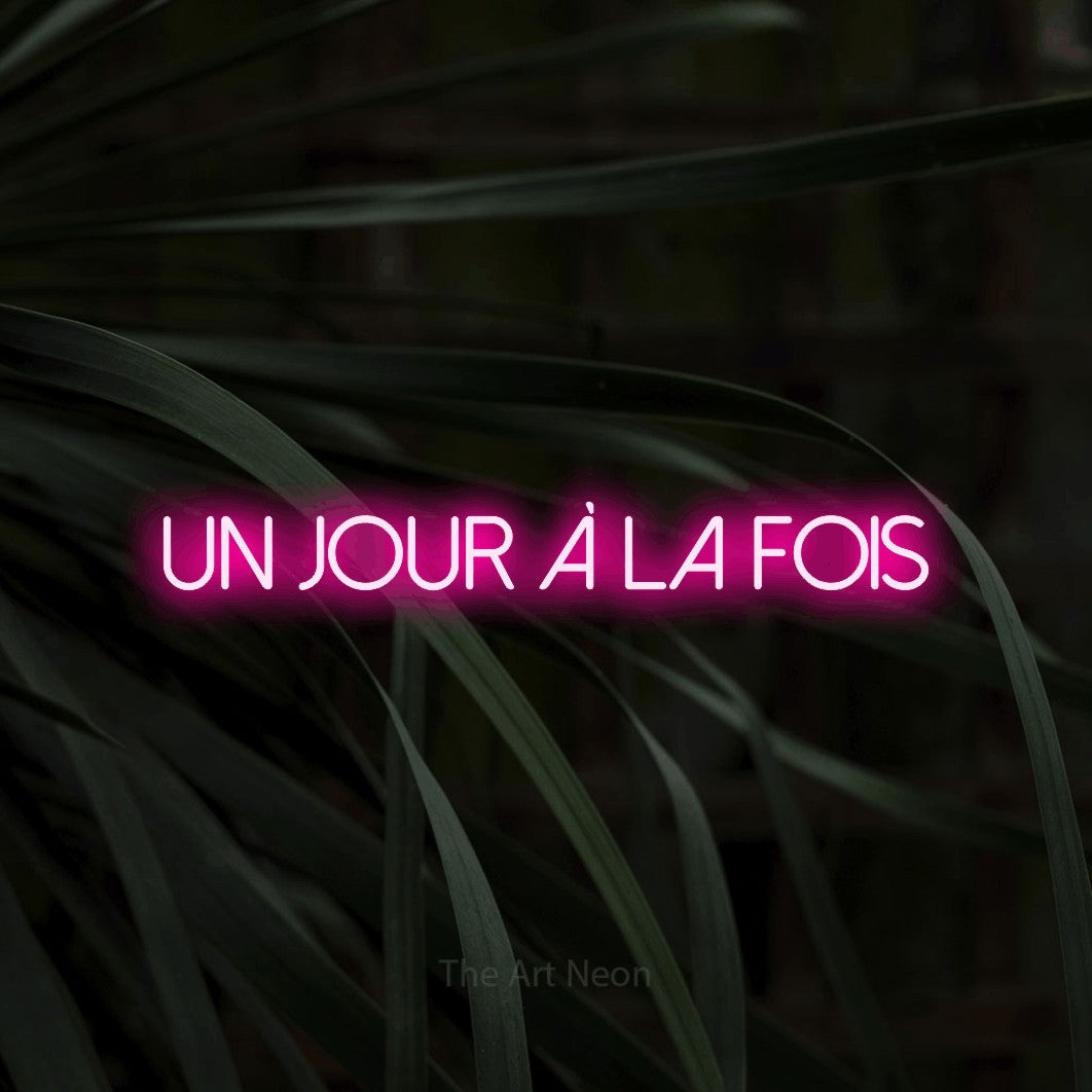 “Un jour à la fois” LED neon sign in hot pink, with thin sans-serif lettering, shown against a close-up of dark foliage.