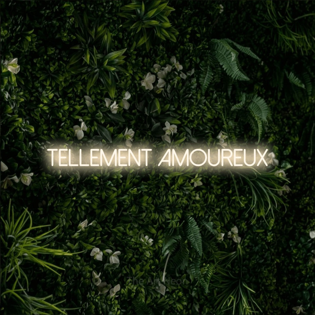 “Tellement amoureux” LED neon sign in warm white, with thin sans-serif lettering, shown against a dense green plant wall.