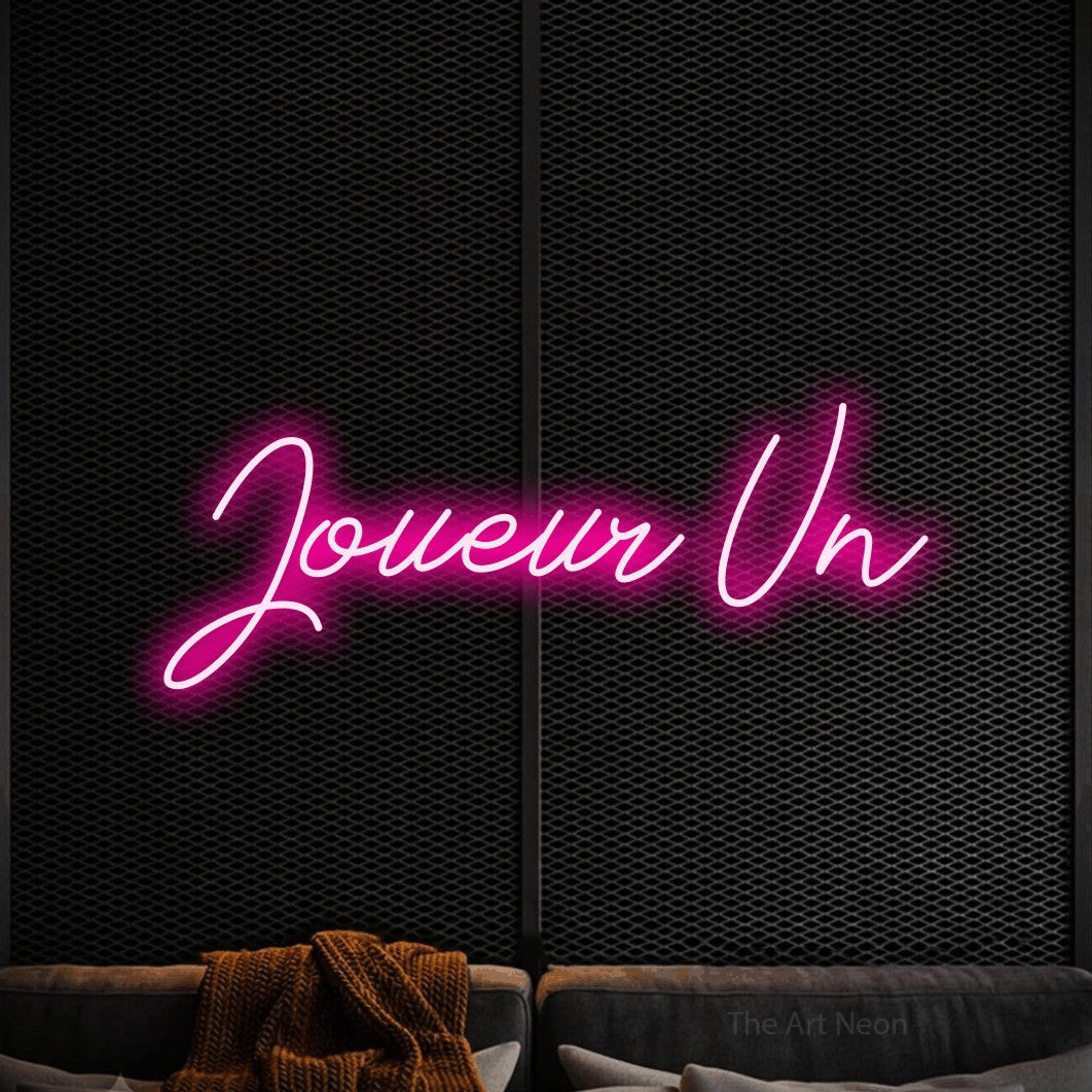 “Joueur un” LED neon sign in hot pink, with cursive lettering, shown against a modern dark-toned seating area.
