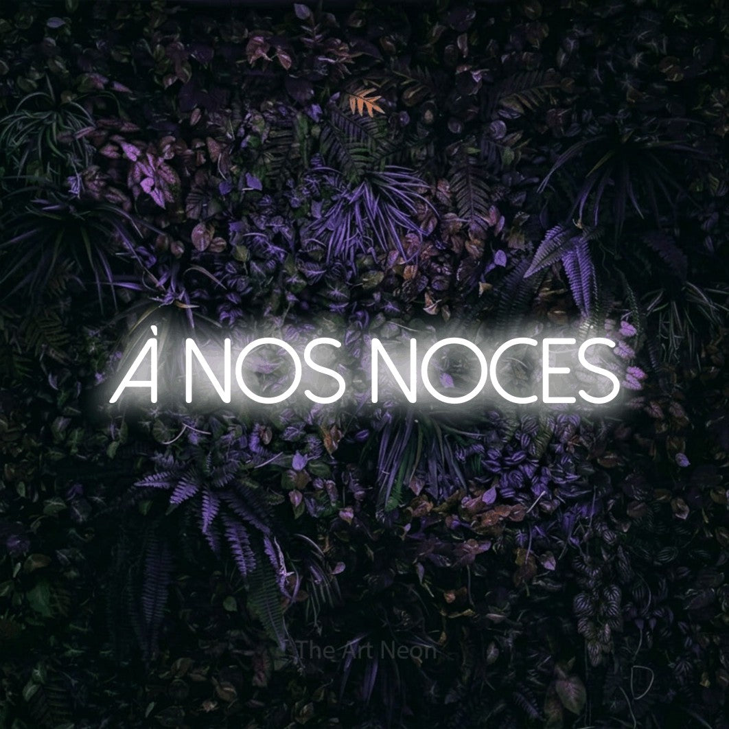 “À nos noces” LED neon sign in cool white, with thin sans-serif lettering, shown against a dark wall with purple flowers.