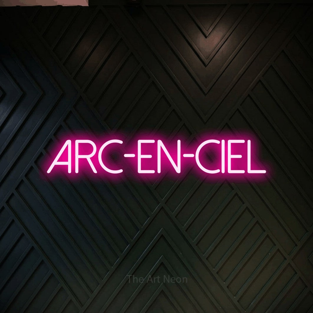 “Arc-en-ciel” LED neon sign in hot pink, with thin sans-serif lettering, shown against a black chevron-patterned wood wall.