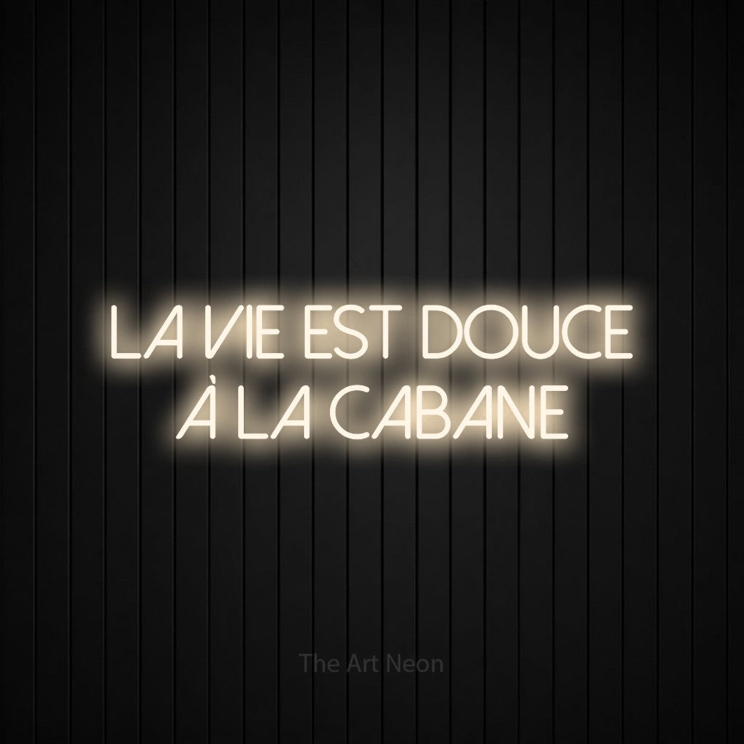 “La vie est douce à la cabane” LED neon sign in warm white, with thin sans-serif lettering across 2 lines, shown against black vertical wall slats.