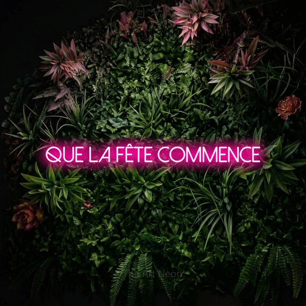 “Que la fête commence” LED neon sign in hot pink, with thin sans-serif lettering, shown against a flowering plant wall.