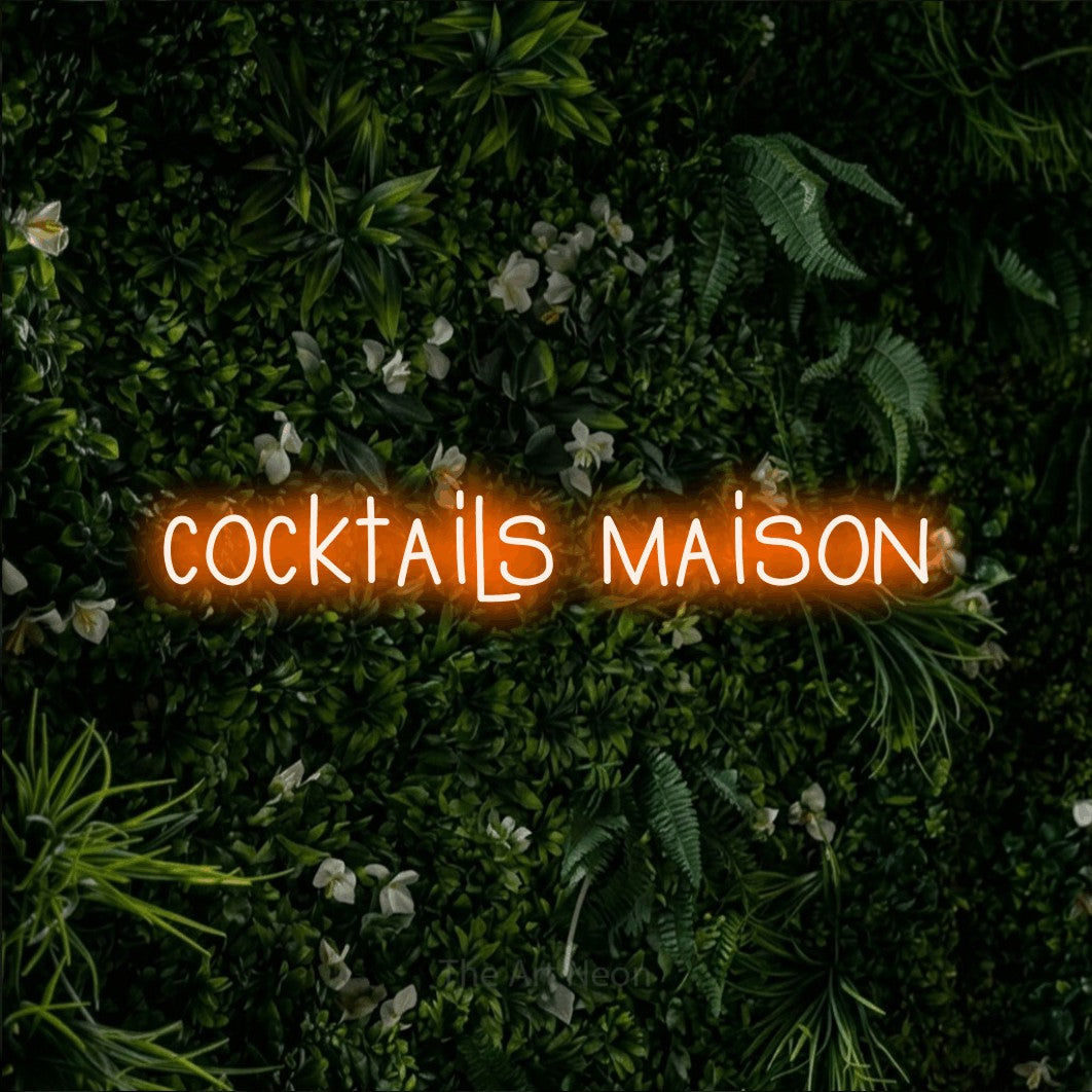 “Cocktails maison” LED neon sign in orange, with narrow hand-drawn lettering, shown against a dense green plant wall.