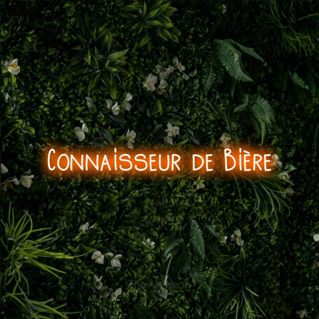 “Connaisseur de bière” LED neon sign in orange, with narrow hand-drawn lettering, shown against a dense green plant wall.