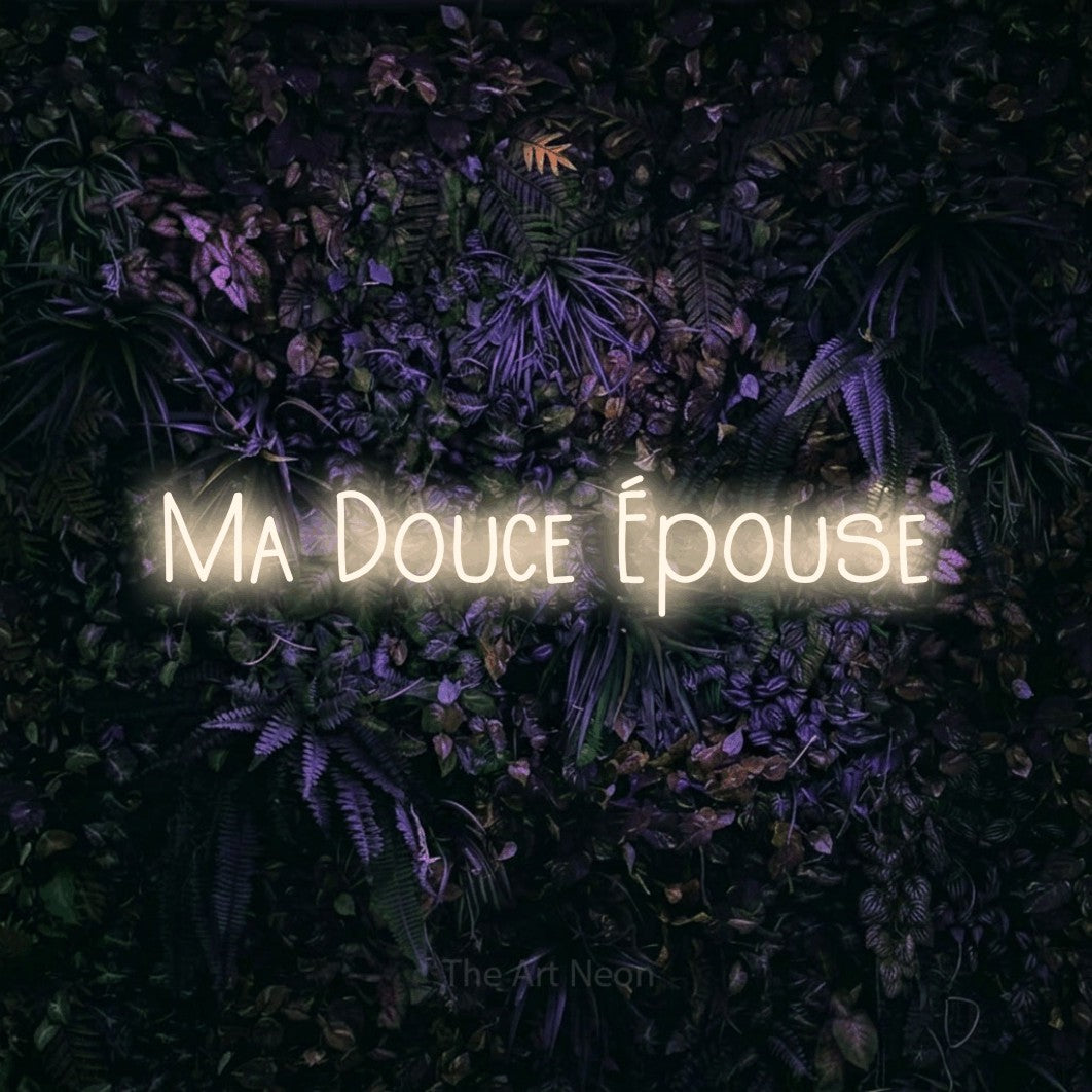 “Ma douce épouse” LED neon sign in warm white, with narrow hand-drawn lettering, shown against a dark wall with purple flowers.