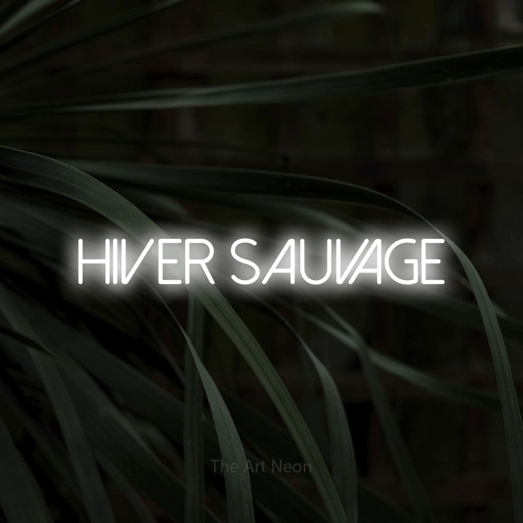 “Hiver sauvage” LED neon sign in cool white, with thin sans-serif lettering, shown against a close-up of dark foliage.