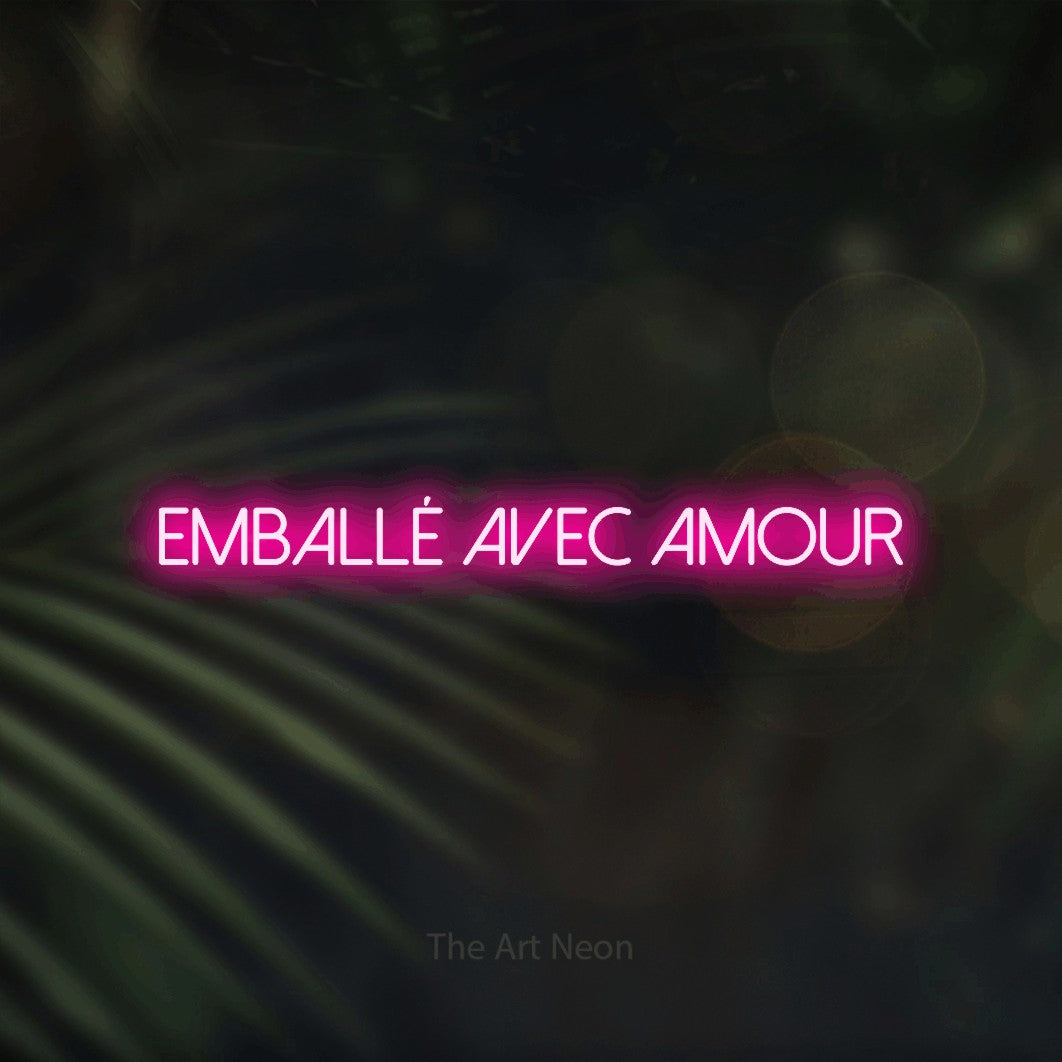 “Emballé avec amour” LED neon sign in hot pink, with thin sans-serif lettering, shown against a blurred palm and bokeh-light backdrop.
