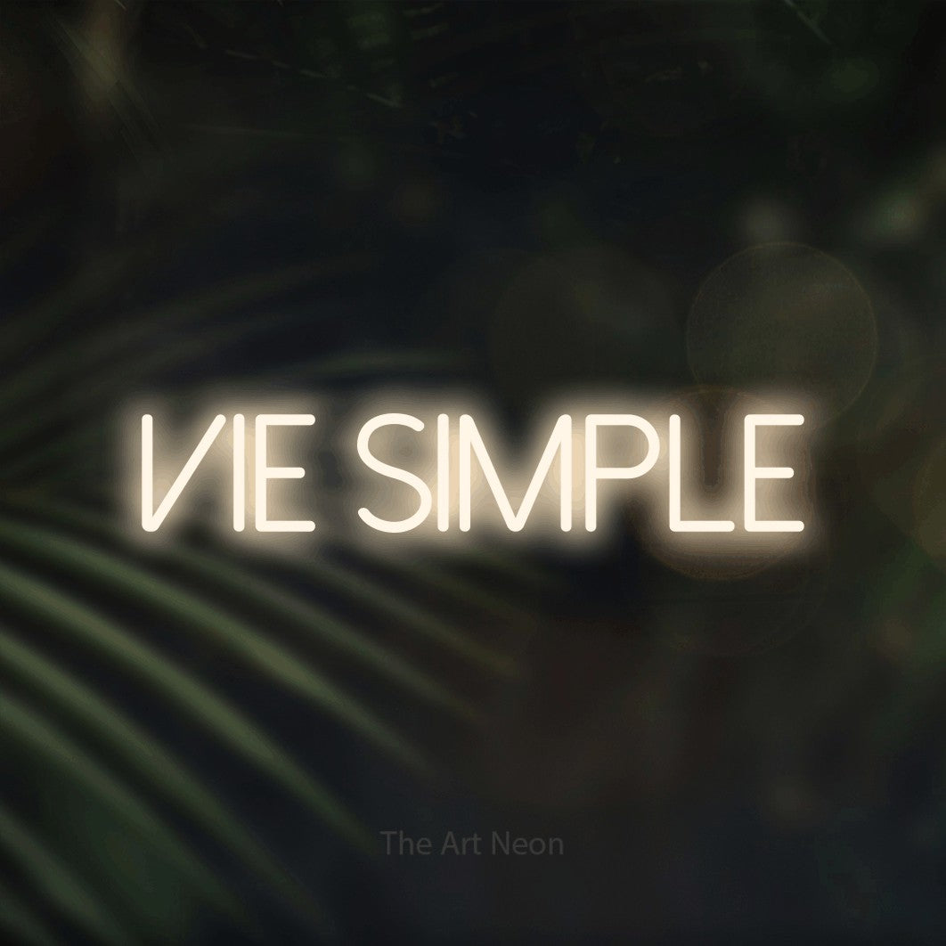 “Vie simple” LED neon sign in warm white, with thin sans-serif lettering, shown against a blurred palm and bokeh-light backdrop.