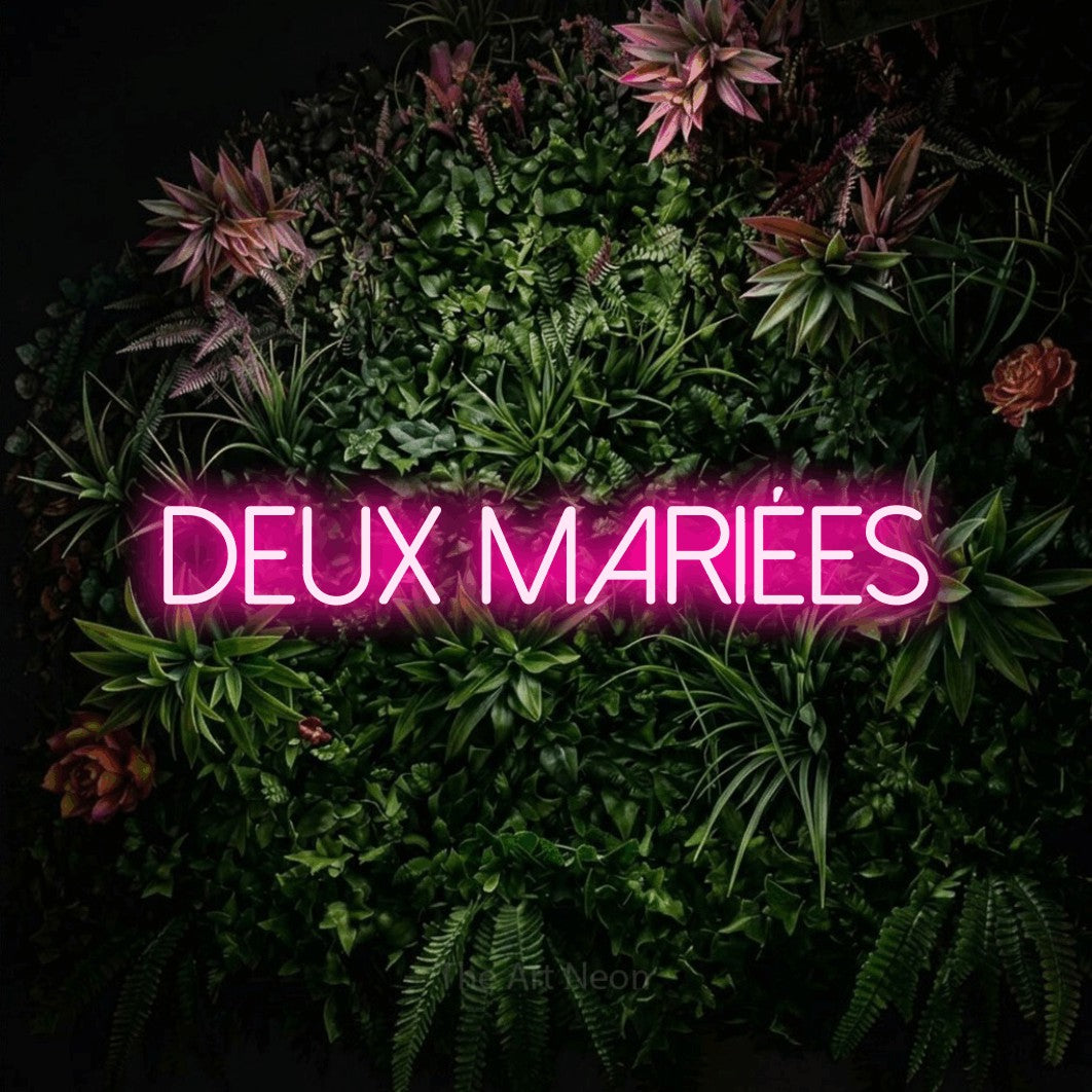 “Deux mariées” LED neon sign in hot pink, with thin sans-serif lettering, shown against a flowering plant wall.
