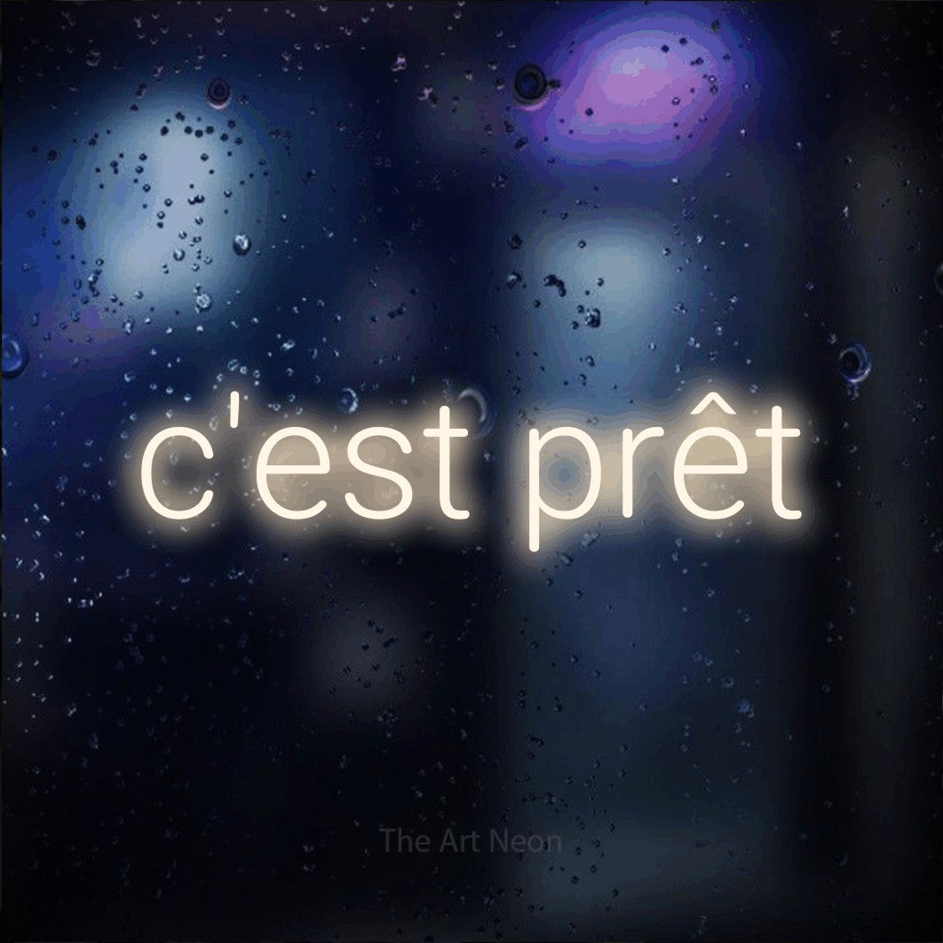 “C'est prêt” LED neon sign in warm white, with rounded sans-serif lettering, shown against glass with blue and purple reflections.
