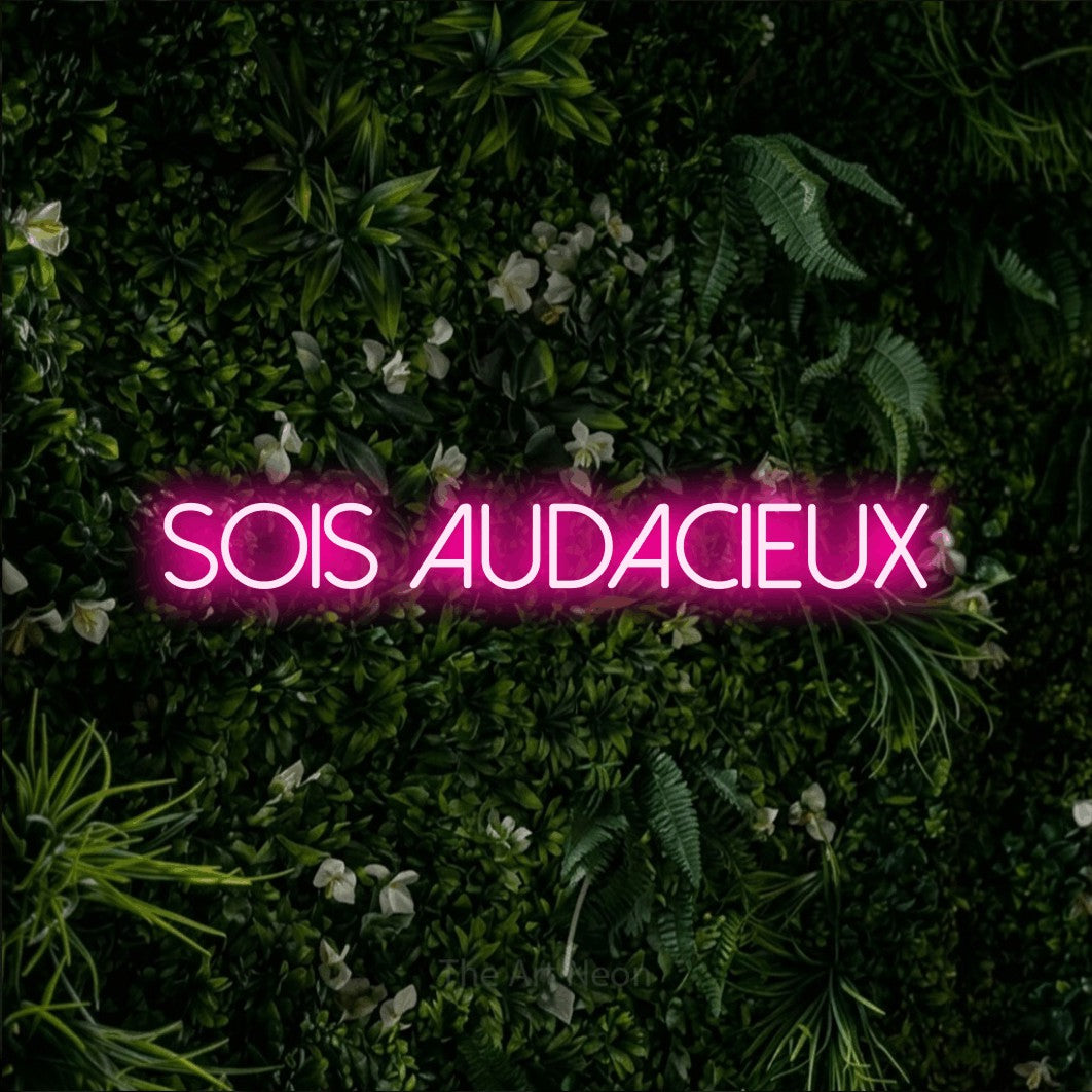“Sois audacieux” LED neon sign in hot pink, with thin sans-serif lettering, shown against a dense green plant wall.