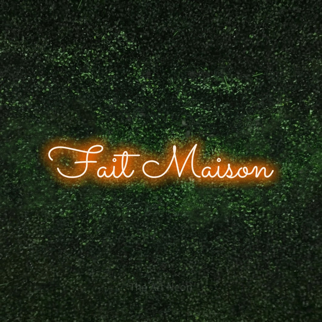 “Fait maison” LED neon sign in orange, with looped cursive lettering, shown against a textured green hedge.