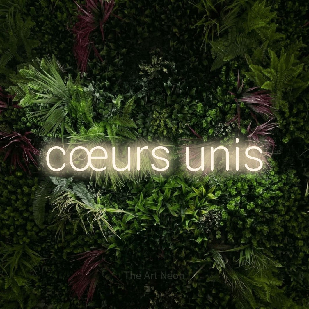 Lettrage en néon LED « Cœurs unis » en blanc chaud, en lettres sans empattement arrondies, présenté sur un mur végétal aux fleurs roses, pour une réception de mariage.
