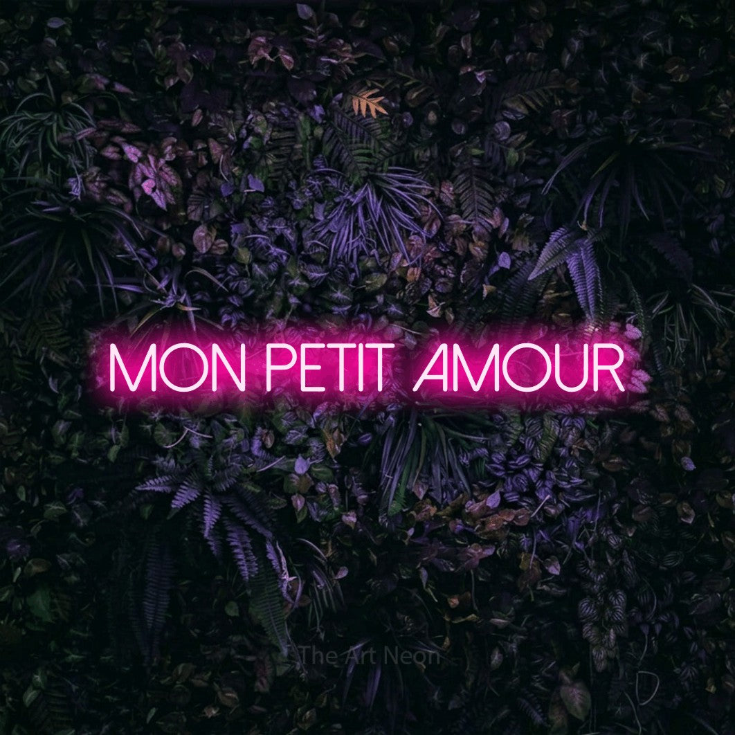 Aperçu d’une enseigne néon LED « Mon petit amour » en rose vif, en lettres fines sans empattement, photographiée sur un mur sombre aux fleurs violettes, pour une chambre d’enfant.