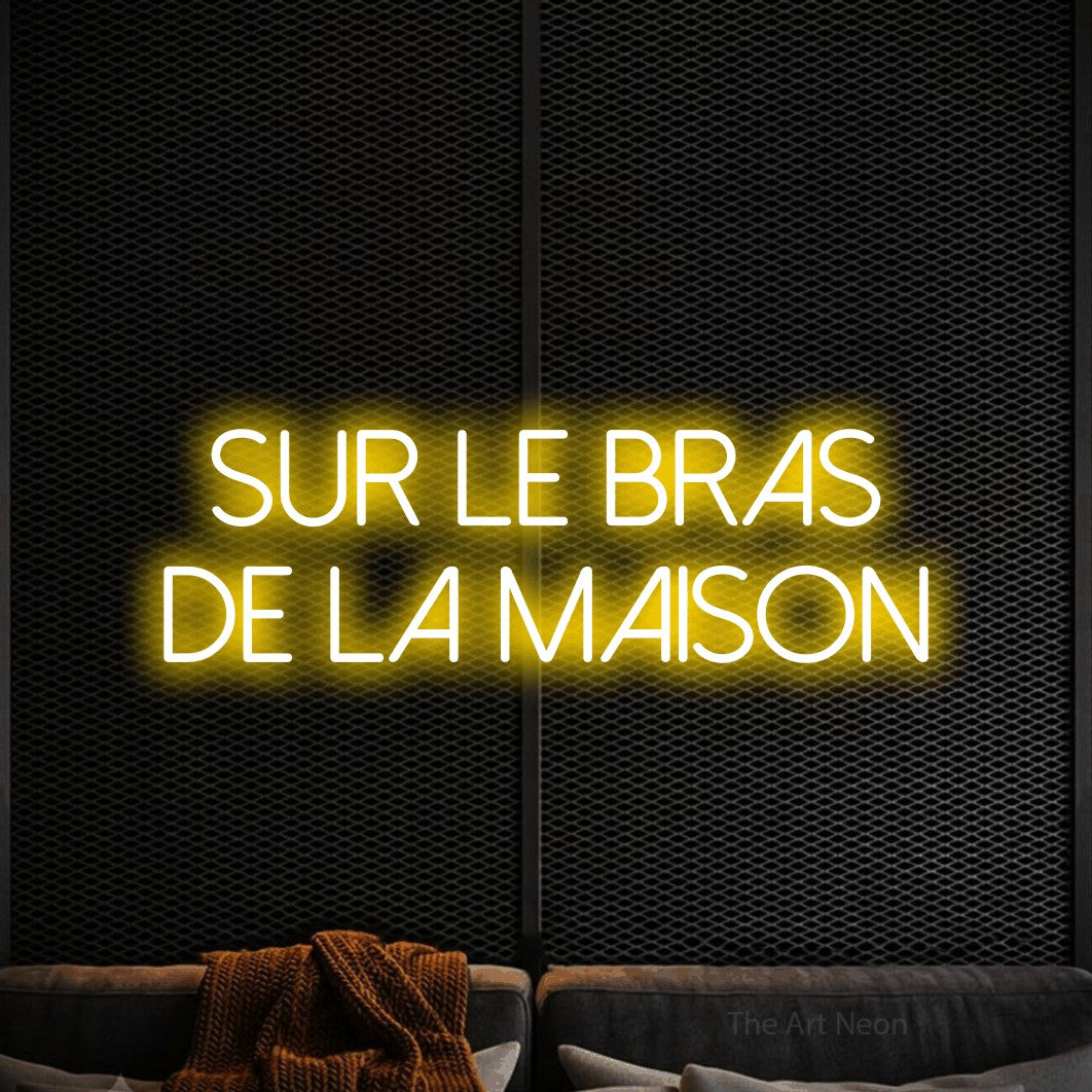 “Sur le bras de la maison” LED neon sign in yellow, with thin sans-serif lettering across 2 lines, shown against a modern dark-toned seating area.