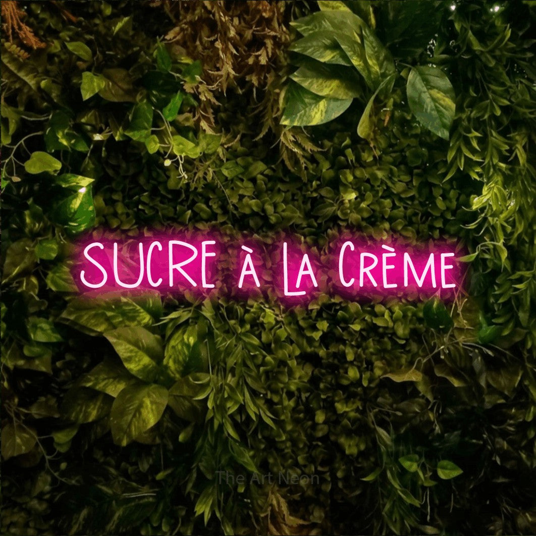 “Sucre à la crème” LED neon sign in hot pink, with narrow hand-drawn lettering, shown against bright green and yellow foliage.