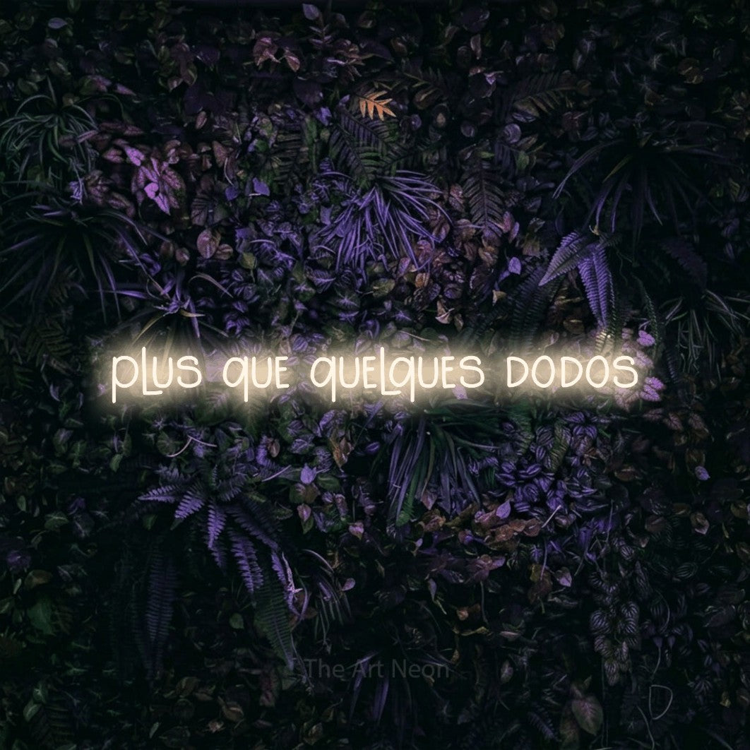 Composition en néon LED « Plus que quelques dodos » en blanc chaud, en lettres étroites dessinées à la main, mise en valeur sur un mur sombre aux fleurs violettes, pour une fête prénatale.