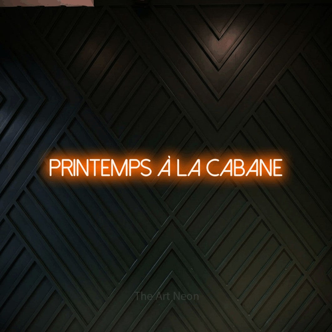 “Printemps à la cabane” LED neon sign in orange, with thin sans-serif lettering, shown against a black chevron-patterned wood wall.