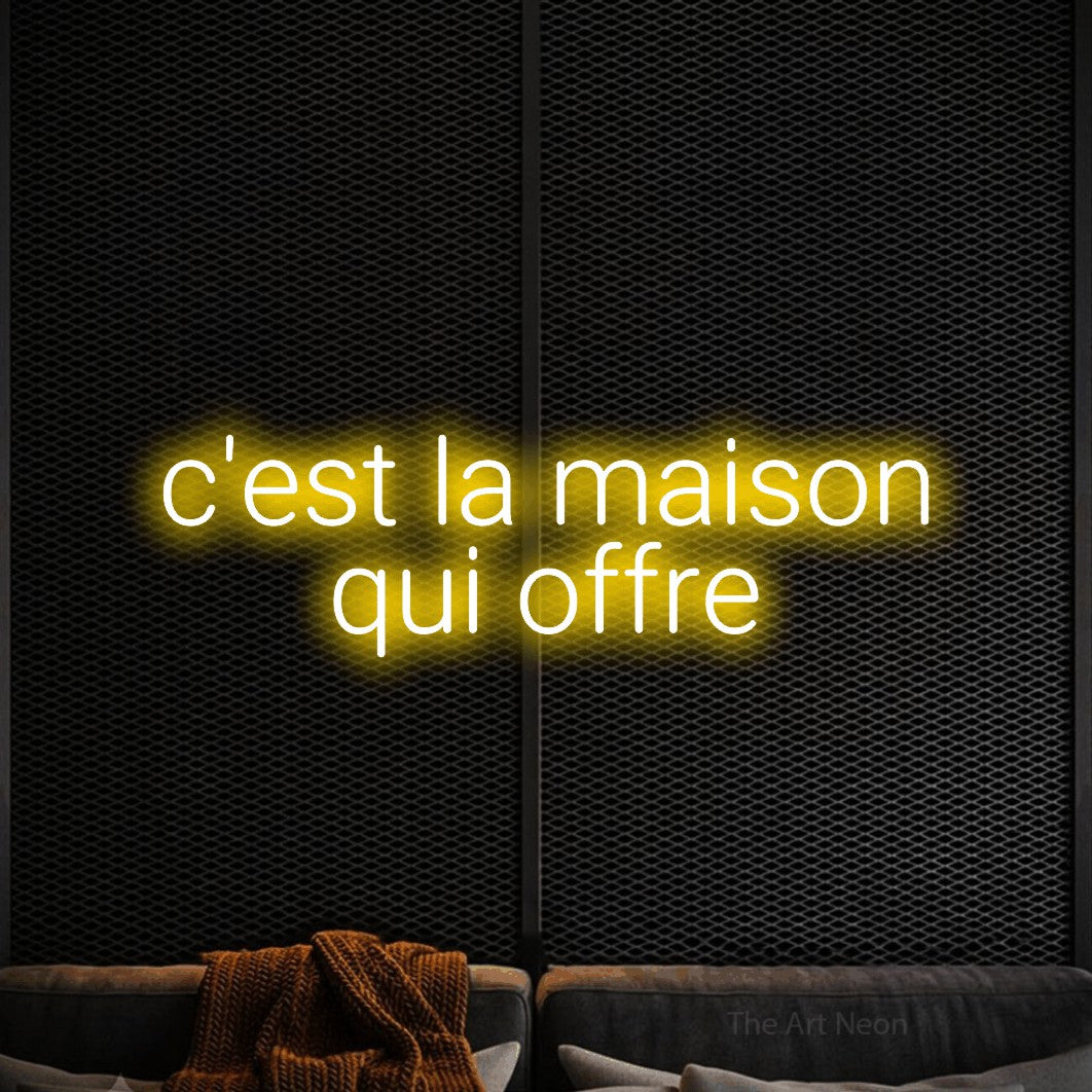 “C'est la maison qui offre” LED neon sign in yellow, with rounded sans-serif lettering across 2 lines, shown against a modern dark-toned seating area.