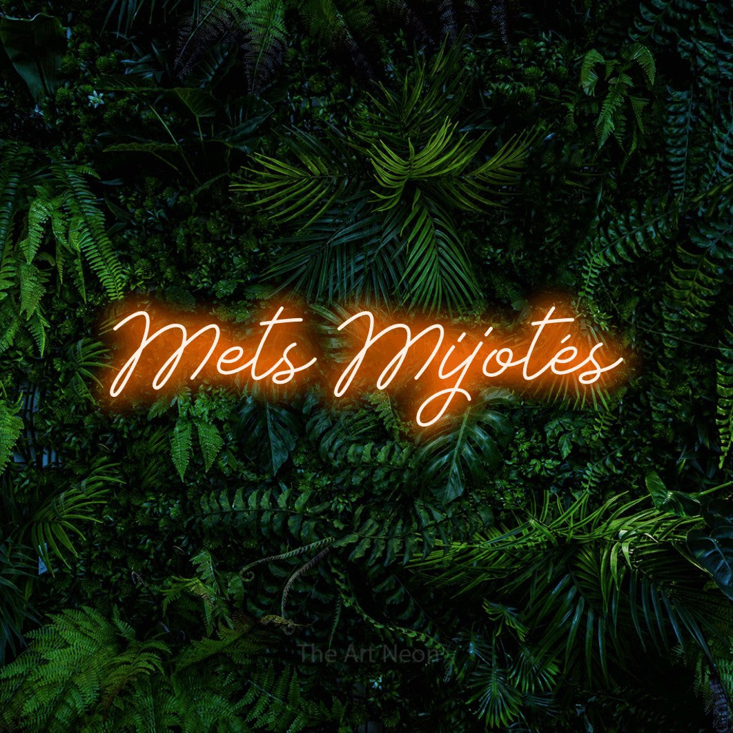 “Mets mijotés” LED neon sign in orange, with cursive lettering, shown against dense tropical foliage.