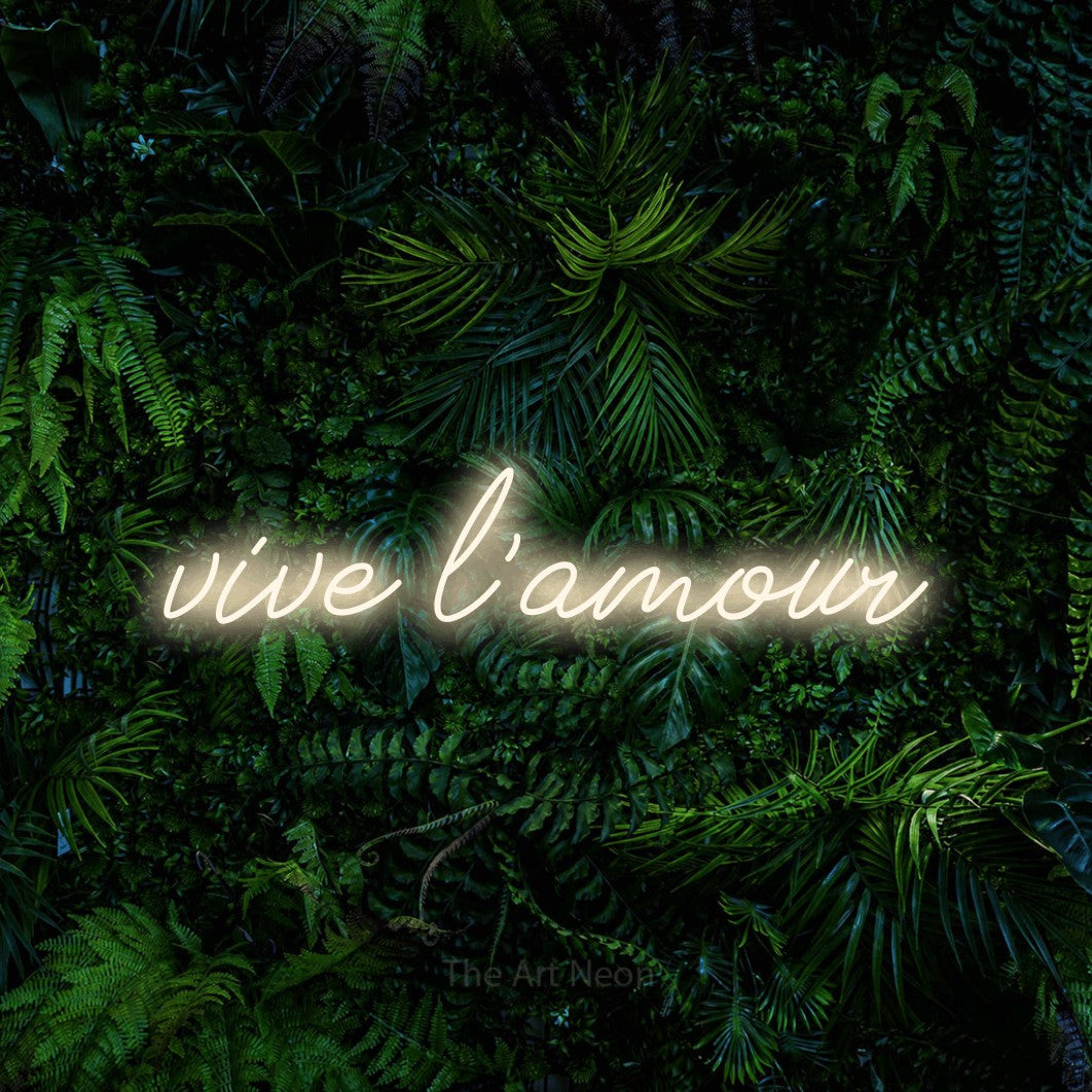 “Vive l'amour” LED neon sign in warm white, with cursive lettering, shown against dense tropical foliage.
