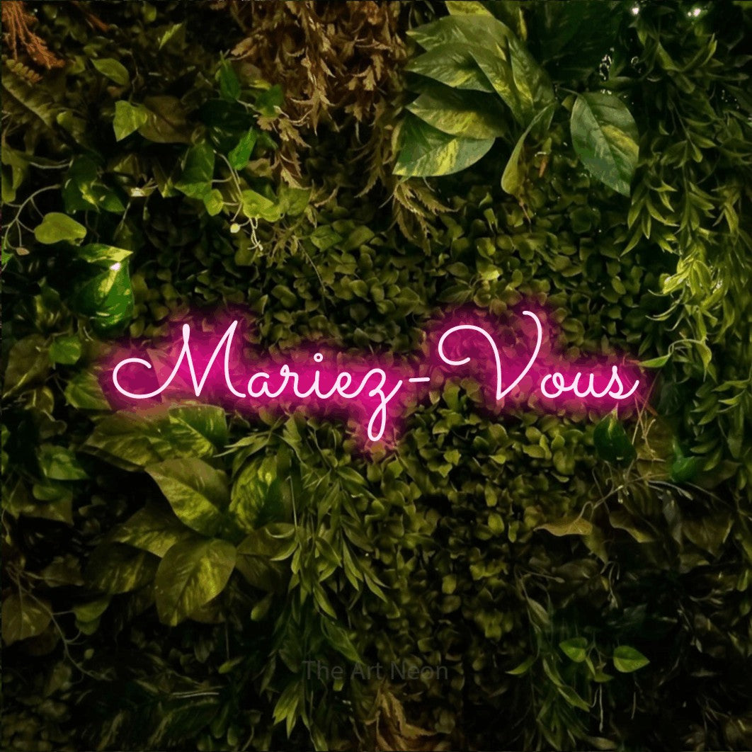 Néon LED « Mariez-vous » en rose vif, en lettres cursives aux boucles marquées, mis en scène sur un feuillage vert et jaune vif, pour une réception de mariage.