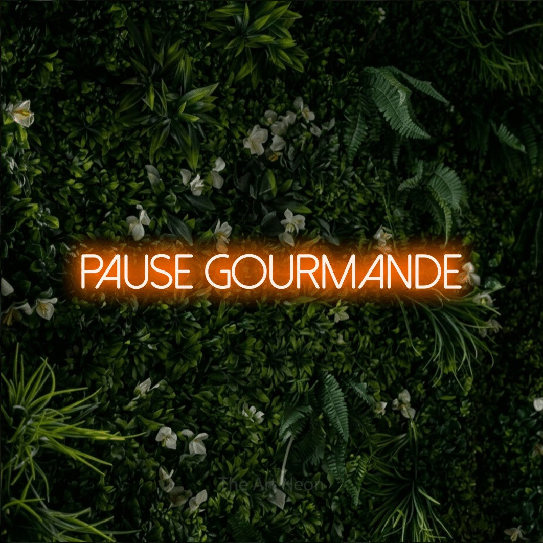 “Pause gourmande” LED neon sign in orange, with thin sans-serif lettering, shown against a dense green plant wall.