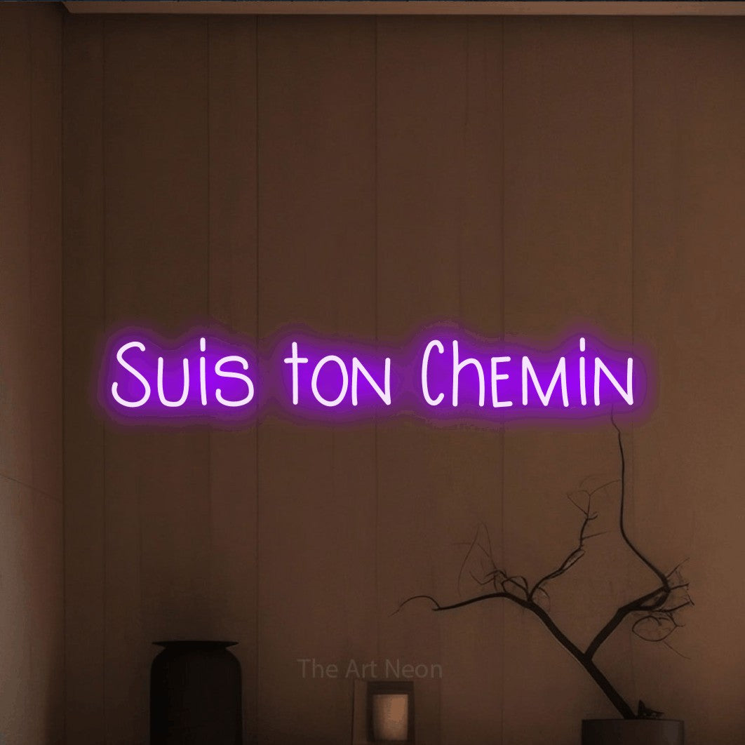 “Suis ton chemin” LED neon sign in purple, with narrow hand-drawn lettering, shown against a simple earth-toned interior.