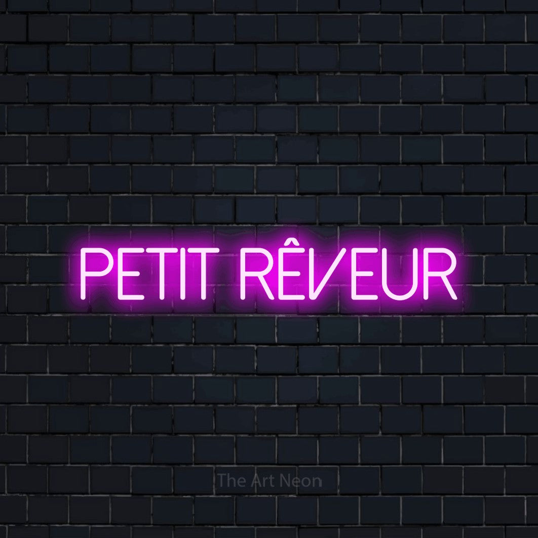 “Petit rêveur” LED neon sign in pink, with thin sans-serif lettering, shown against a dark navy brick wall.