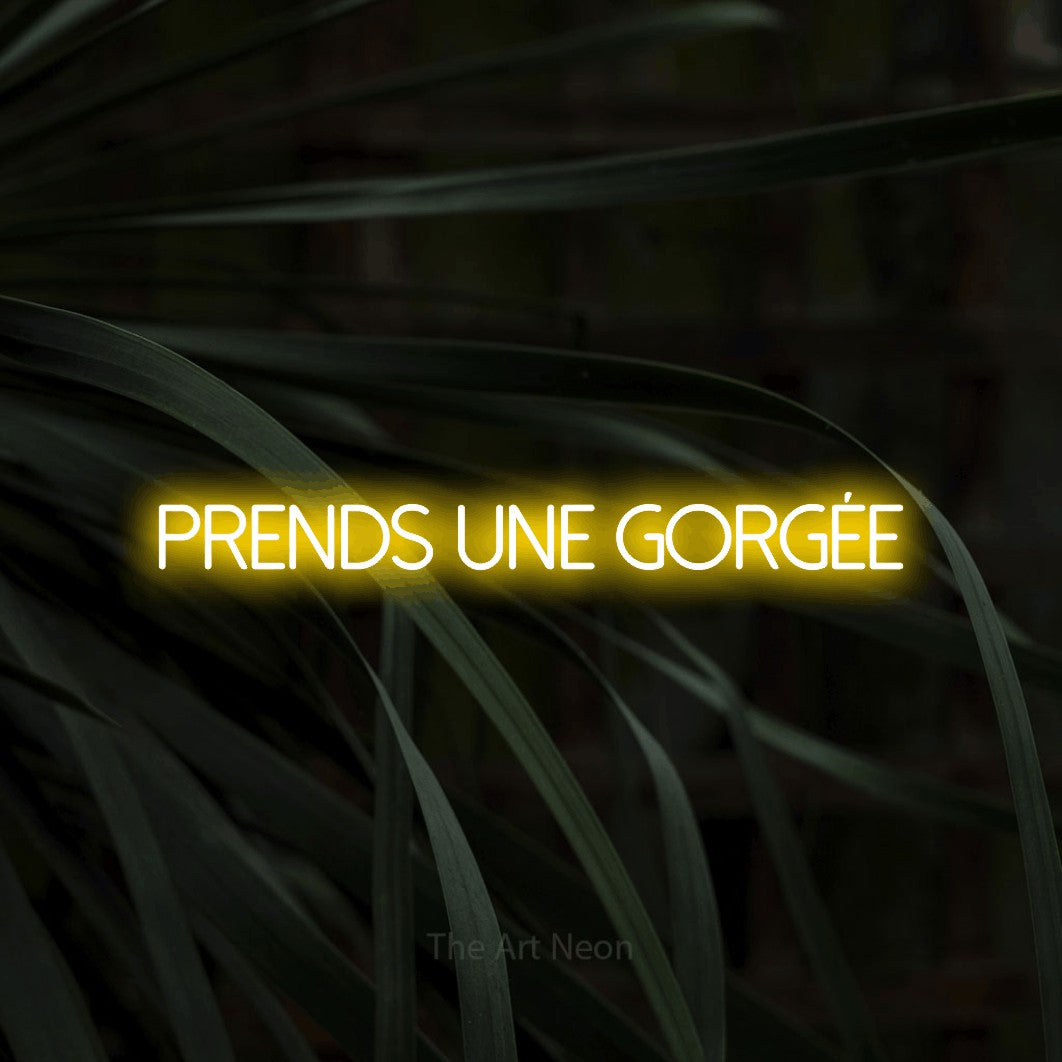 “Prends une gorgée” LED neon sign in yellow, with thin sans-serif lettering, shown against a close-up of dark foliage.
