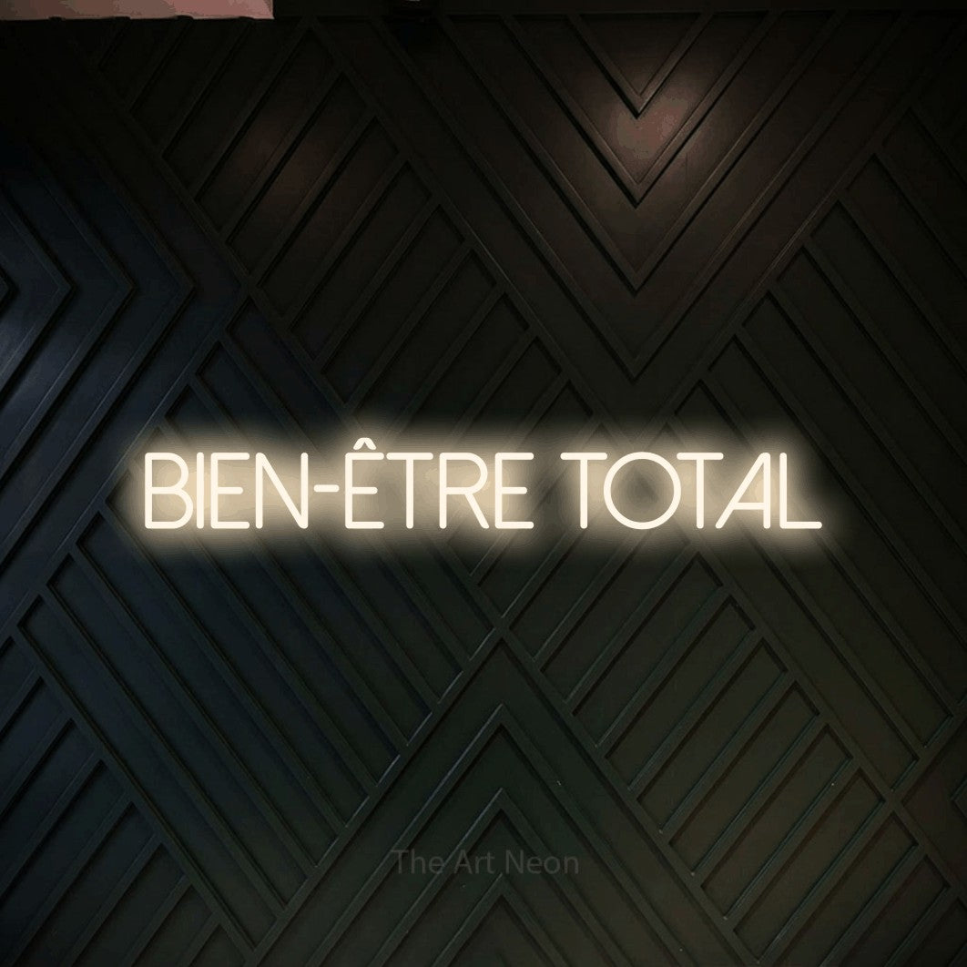 “Bien-être total” LED neon sign in warm white, with thin sans-serif lettering, shown against a black chevron-patterned wood wall.