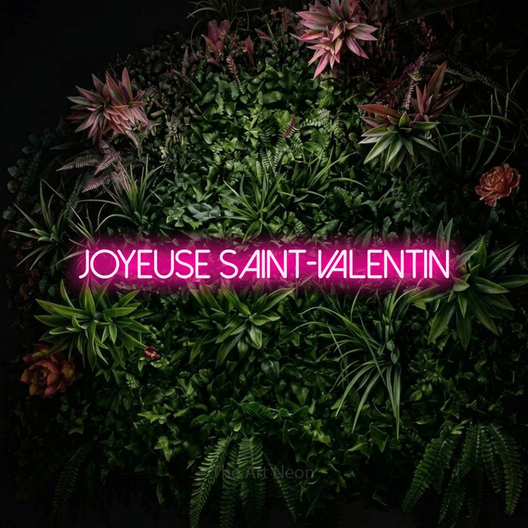“Joyeuse Saint-Valentin” LED neon sign in hot pink, with thin sans-serif lettering, shown against a flowering plant wall.