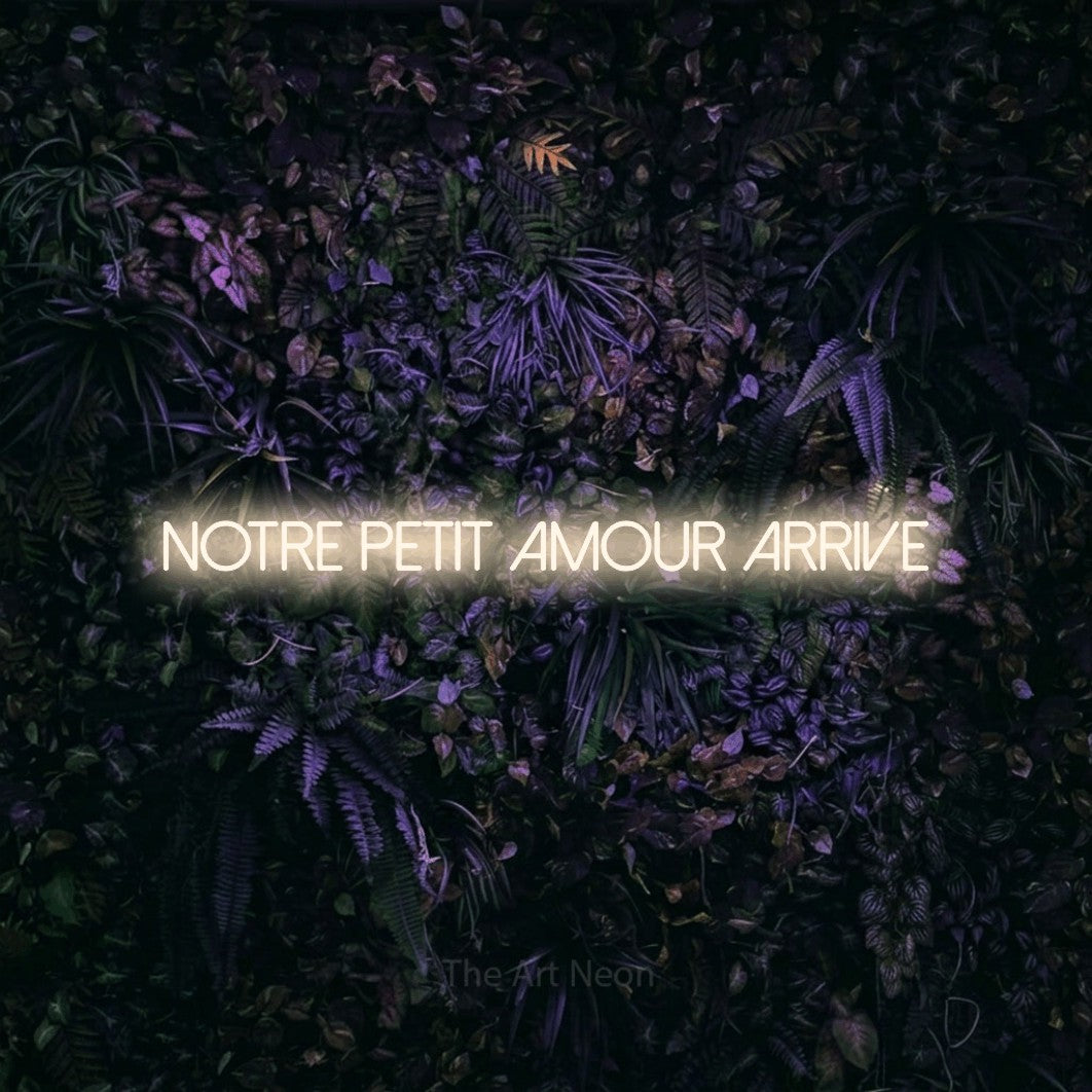 Composition en néon LED « Notre petit amour arrive » en blanc chaud, en lettres fines sans empattement, mise en valeur sur un mur sombre aux fleurs violettes, pour une fête prénatale.