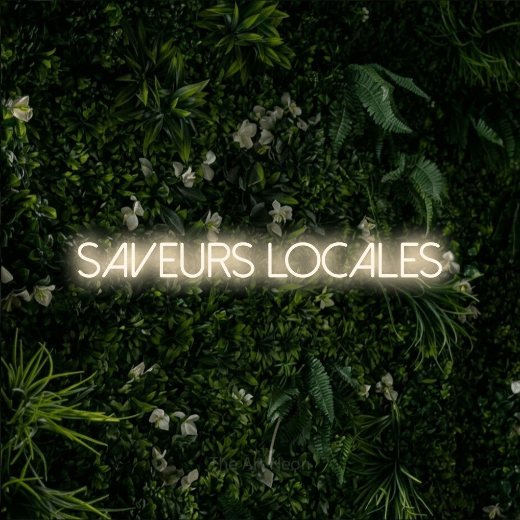 “Saveurs locales” LED neon sign in warm white, with thin sans-serif lettering, shown against a dense green plant wall.