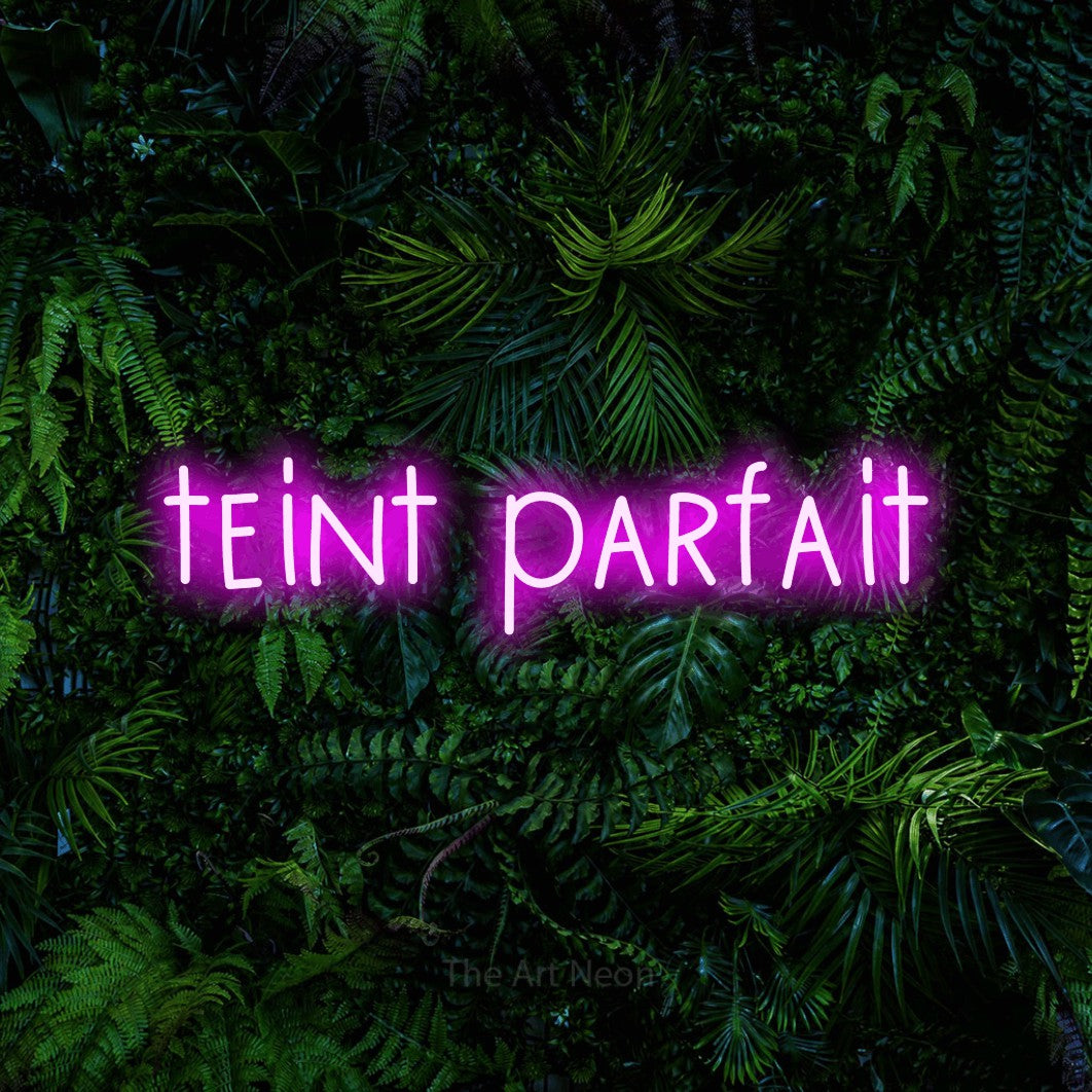 “Teint parfait” LED neon sign in pink, with narrow hand-drawn lettering, shown against dense tropical foliage.