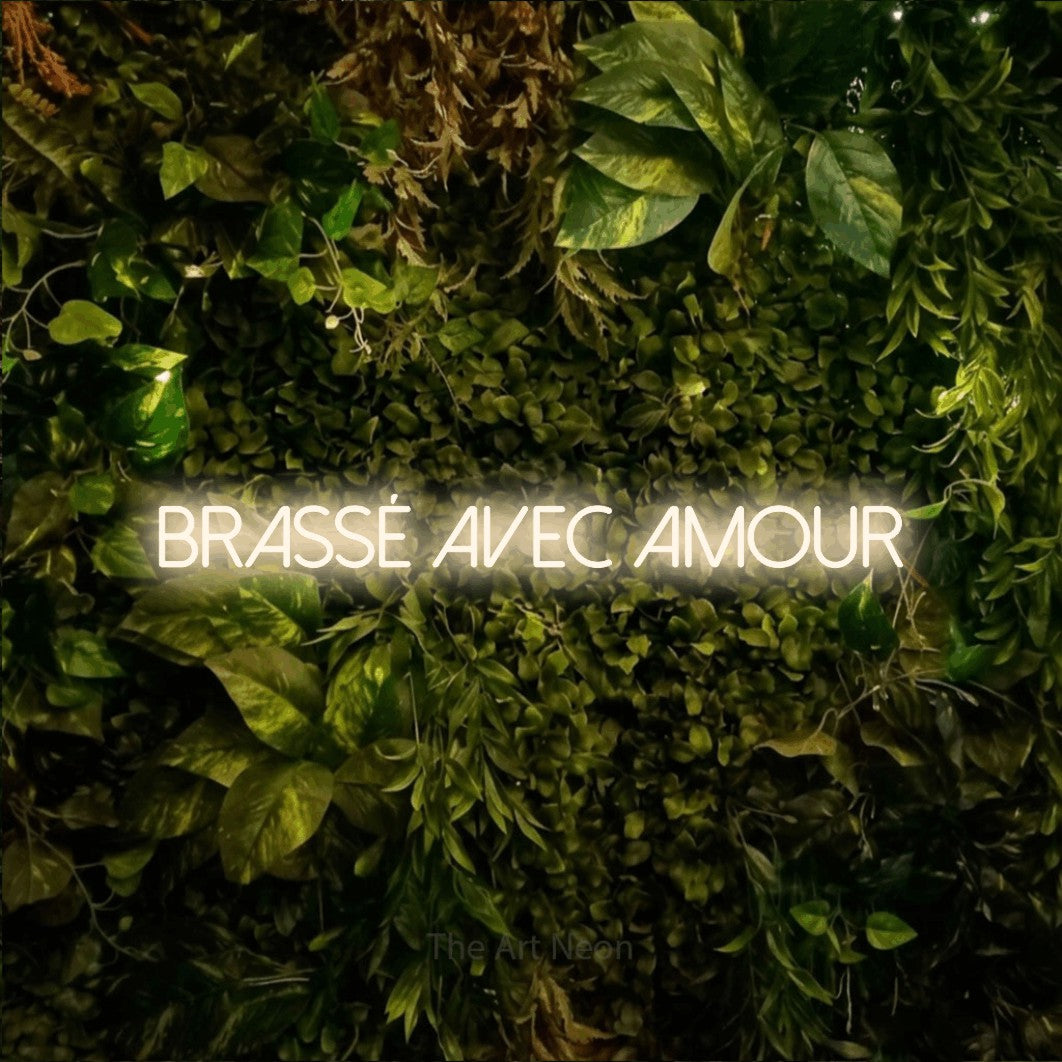 “Brassé avec amour” LED neon sign in warm white, with thin sans-serif lettering, shown against bright green and yellow foliage.