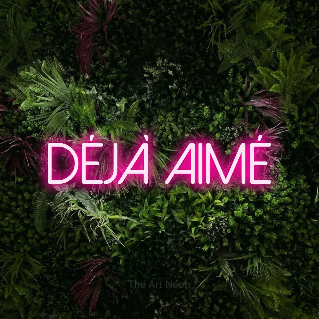 “Déjà aimé” LED neon sign in hot pink, with thin sans-serif lettering, shown against a plant wall with pink flowers.
