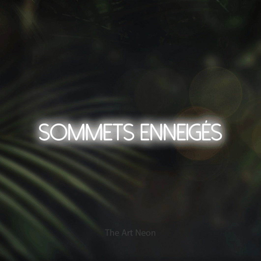 “Sommets enneigés” LED neon sign in cool white, with thin sans-serif lettering, shown against a blurred palm and bokeh-light backdrop.