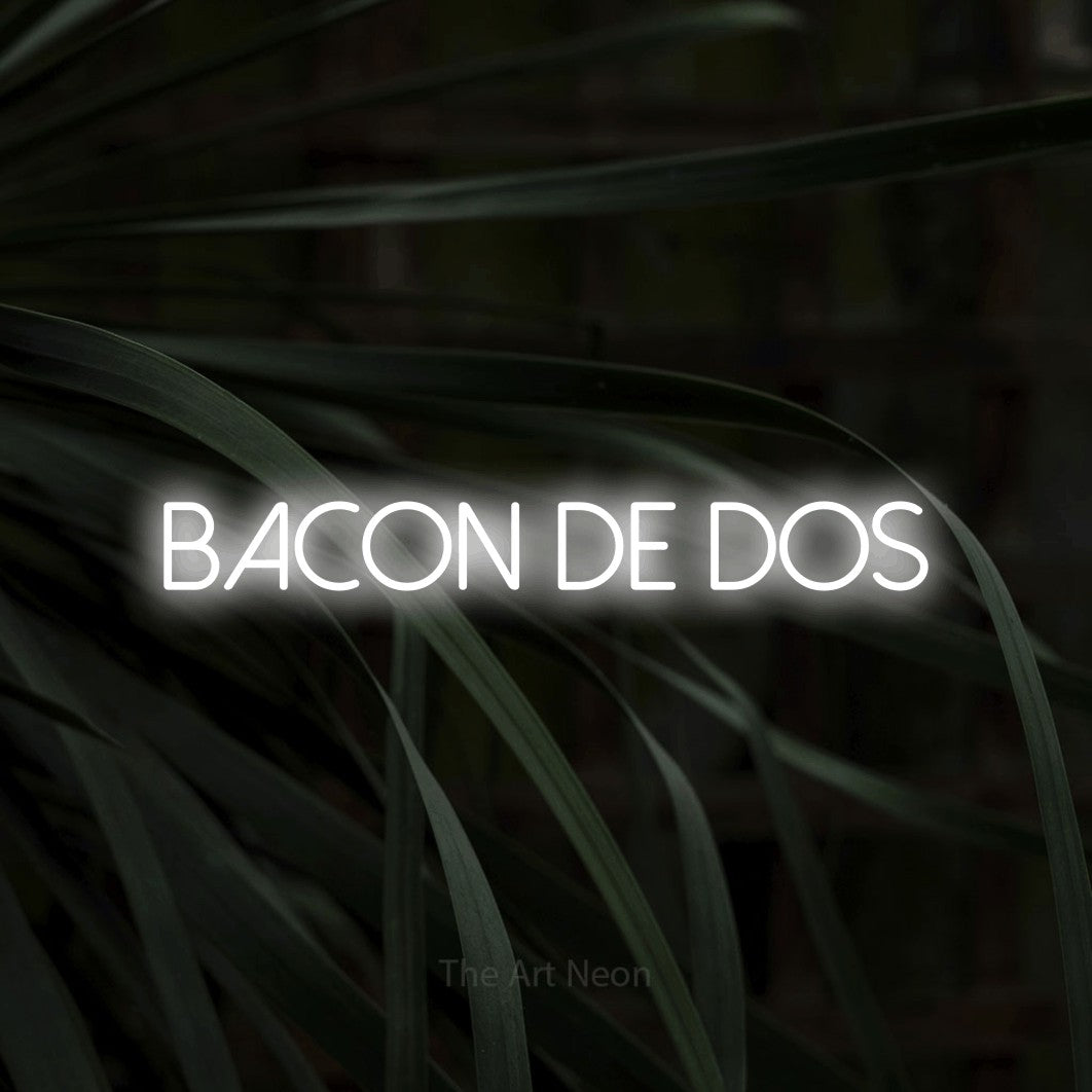“Bacon de dos” LED neon sign in cool white, with thin sans-serif lettering, shown against a close-up of dark foliage.