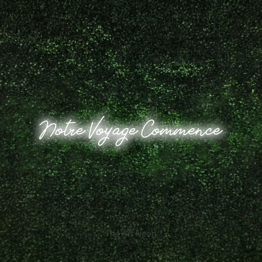 “Notre voyage commence” LED neon sign in cool white, with cursive lettering, shown against a textured green hedge.