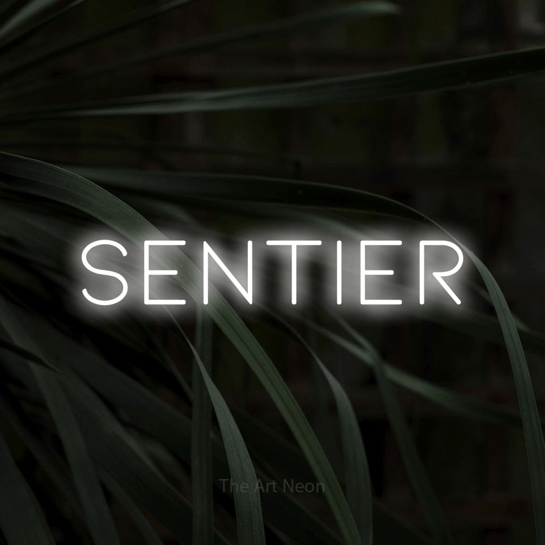 “Sentier” LED neon sign in cool white, with thin, widely spaced lettering, shown against a close-up of dark foliage.