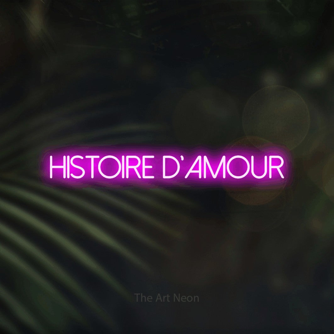 “Histoire d'amour” LED neon sign in pink, with thin sans-serif lettering, shown against a blurred palm and bokeh-light backdrop.