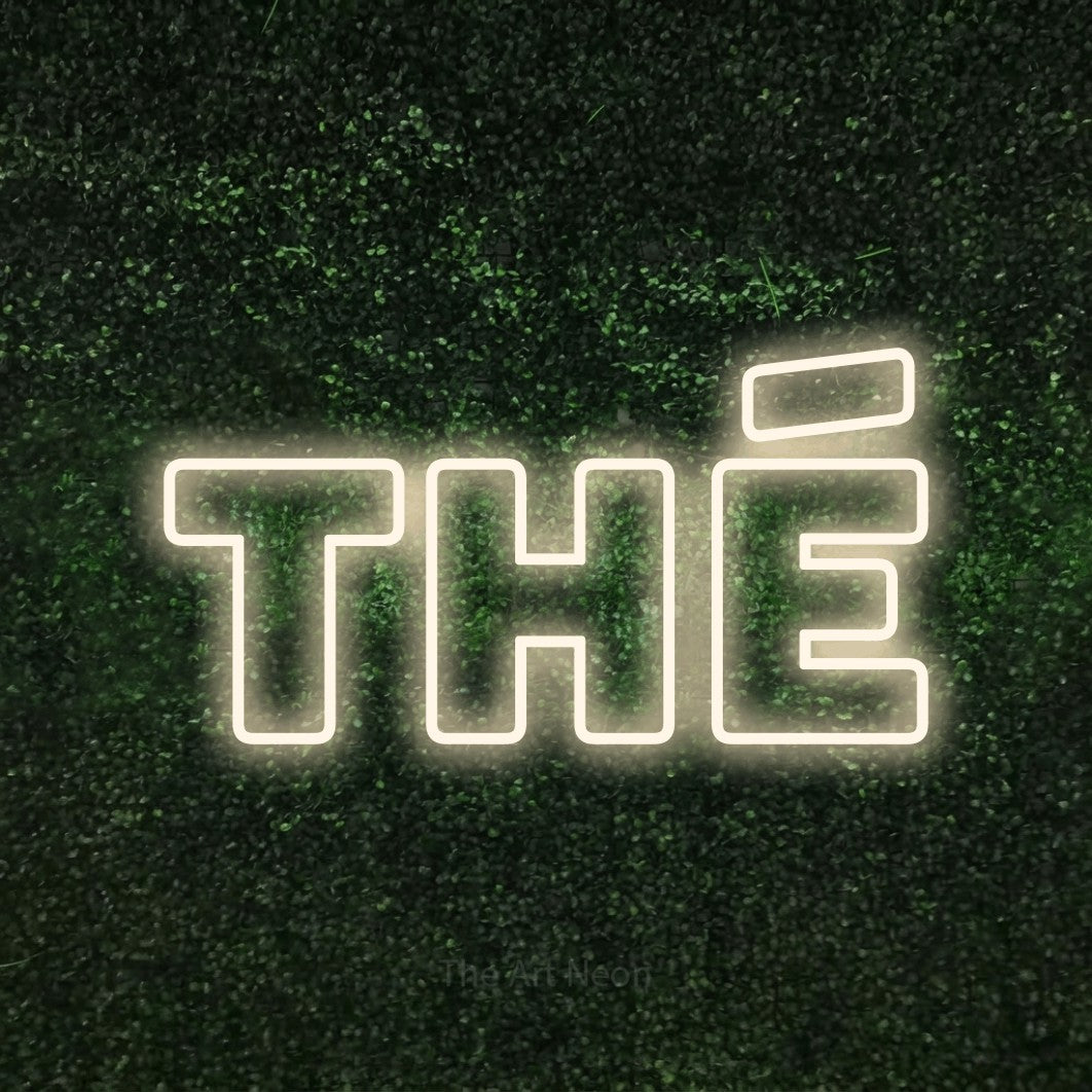“Thé” LED neon sign in warm white, with bold outline lettering, shown against a textured green hedge.