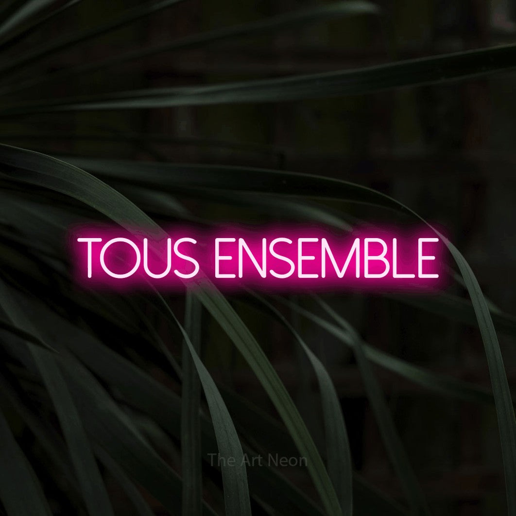 “Tous ensemble” LED neon sign in hot pink, with thin sans-serif lettering, shown against a close-up of dark foliage.